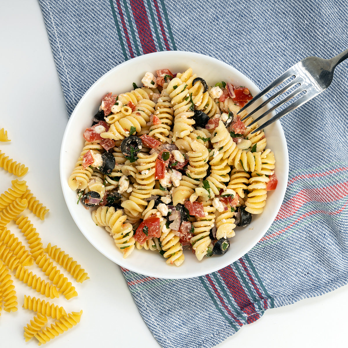 Greek Pasta Salad with rotini, tomato, olives and feta crumbles in a white bowl by Zina's Fine Foods