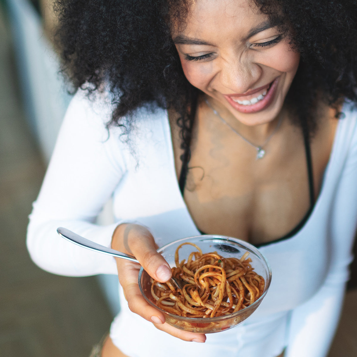 Smiling woman enjoying Zina's Fine Foods Sesame Noodle Salad from a clear glass bowl